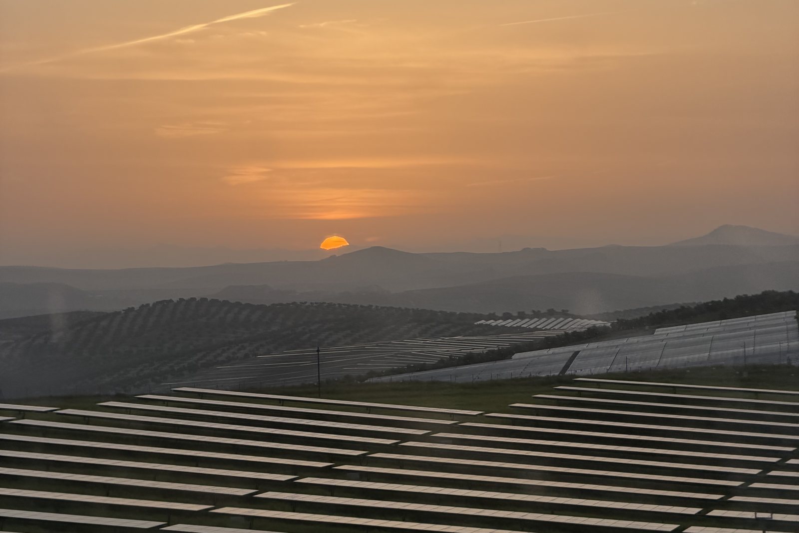 Fotografía de un atardecer sobre una planta fotovoltaica, con el sol ocultándose tras las montañas